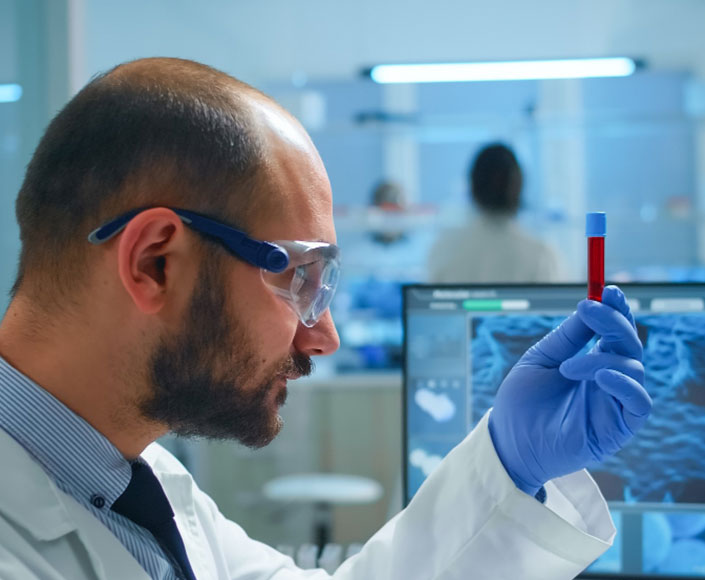 Scientist in safety goggles and blue gloves holds a test tube with red liquid in a lab setting.